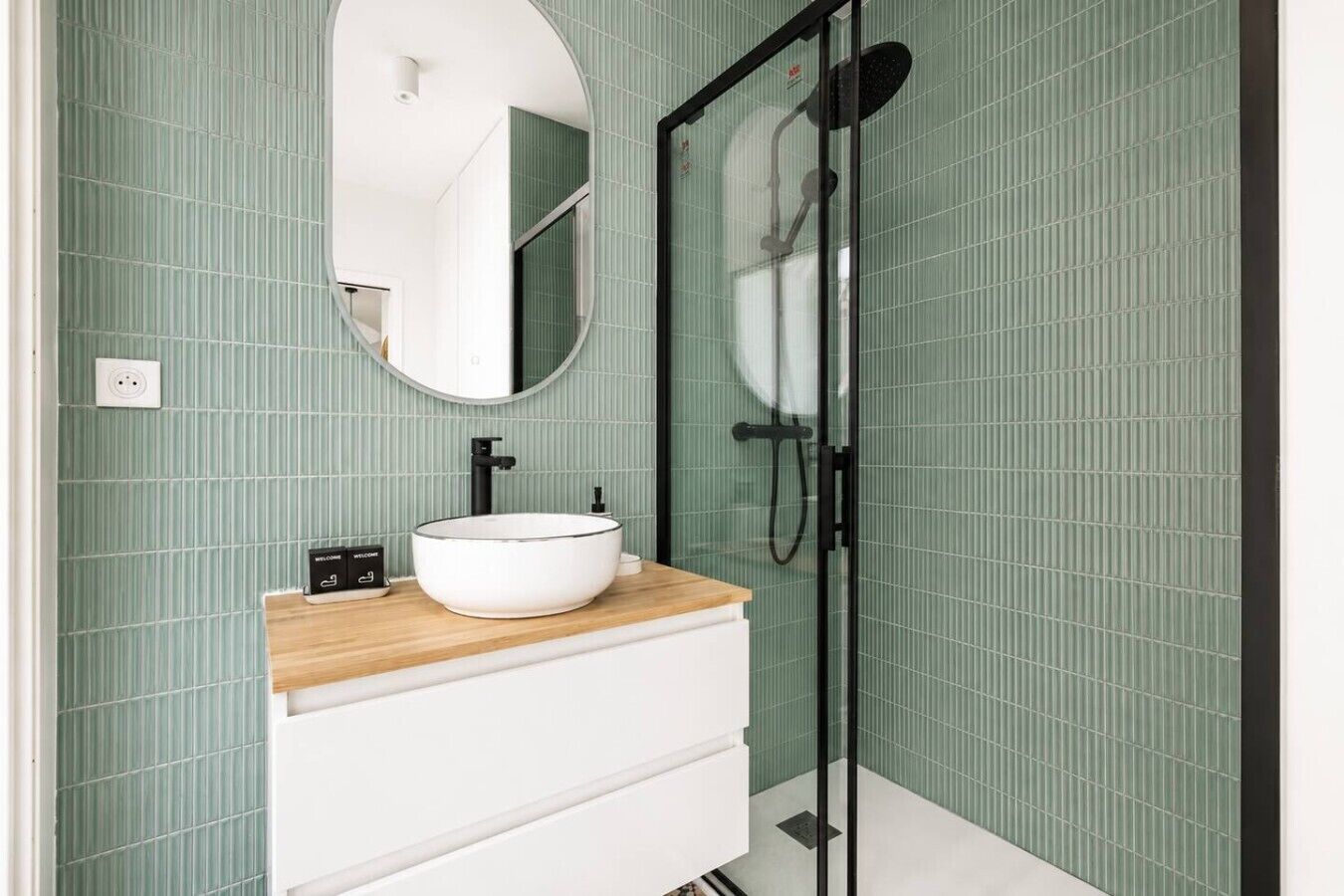 Une salle de bain avec un miroir rond, un lavabo blanc avec un robinet noir et une douche à parois de verre.