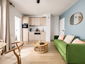 Ein modernes Wohnzimmer mit einem grünen Sofa, einem runden Couchtisch und einer Küchenzeile mit hellen Schränken.