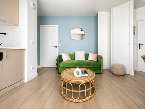 Ein Zimmer mit einem grünen Sofa, einem runden Korbstuhl-Couchtisch, einem Spiegel an der Wand und einer weißen Tür.
