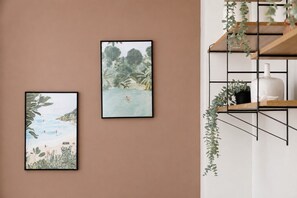Drei gerahmte Kunstwerke an einer Wand, ein Regal mit Pflanzen und eine weiße Vase.