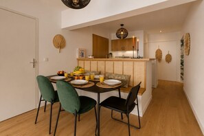 A dining area with a round table, four chairs, and a bench. There is a kitchen in the background with wooden cabinets and a hanging light fixture. - Lavie Maison | Parisian living, Eiffel Tower (Paris)