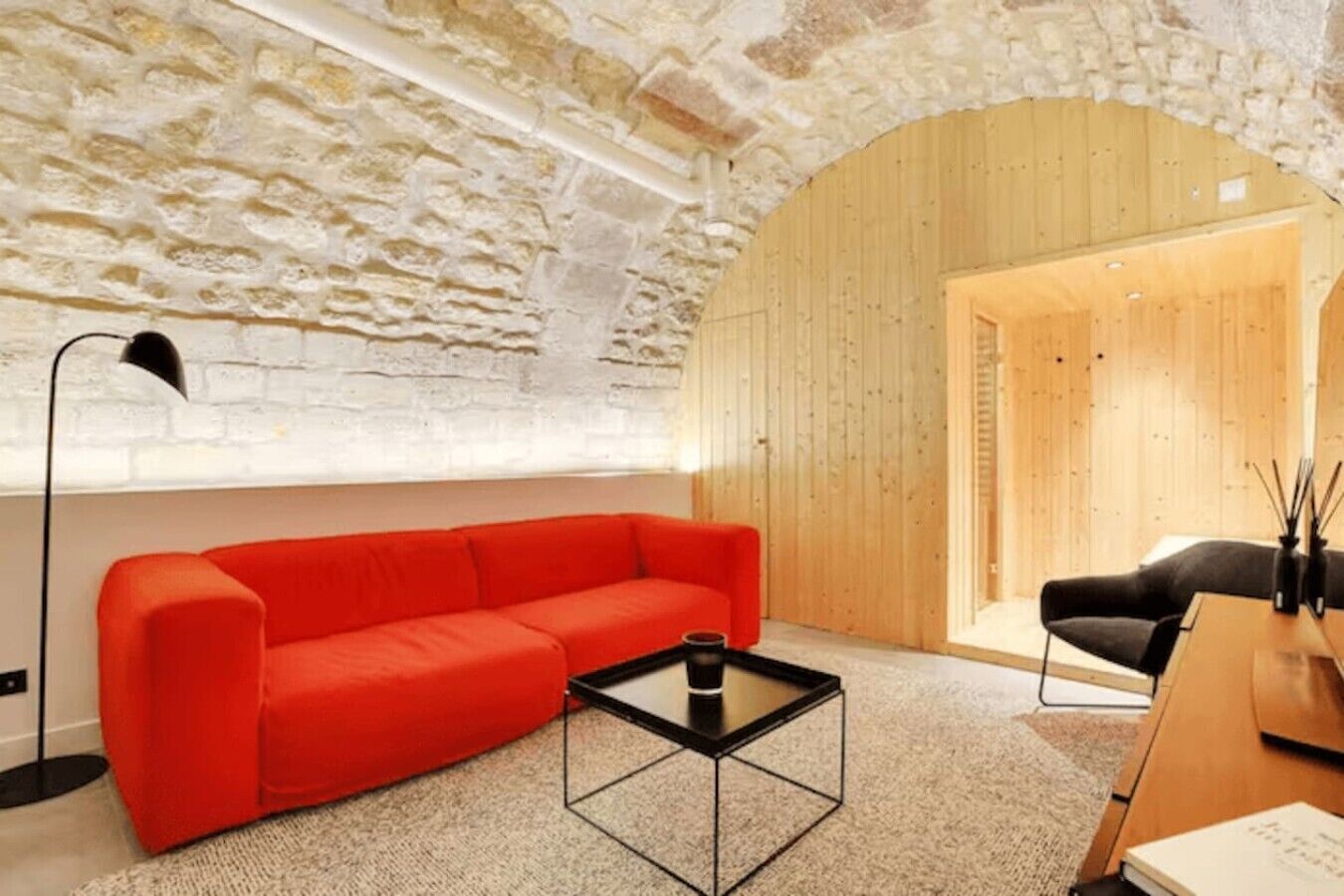 Una habitación con pared de piedra, puerta de madera, sofá rojo, mesa de centro negra y una silla negra.