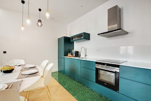 A modern kitchen with teal cabinetry, a stainless steel range hood, and a dining area with white chairs and a table set for two. - Lavie Maison: Private Cinema in Le Marais (Paris)