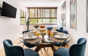 A dining area with a round glass table, blue chairs, and breakfast items like eggs, toast, and fruit. - Lavie Maison | Eiffel Tower & Beaugrenelle (Paris)