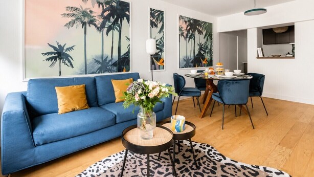 Ein Wohnzimmer mit einem blauen Sofa, einem Couchtisch mit einer Vase sowie einem Essbereich mit Stühlen und Tisch.