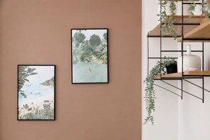 Drei gerahmte Kunstwerke an einer Wand, ein Regal mit Pflanzen und eine weiße Vase.
