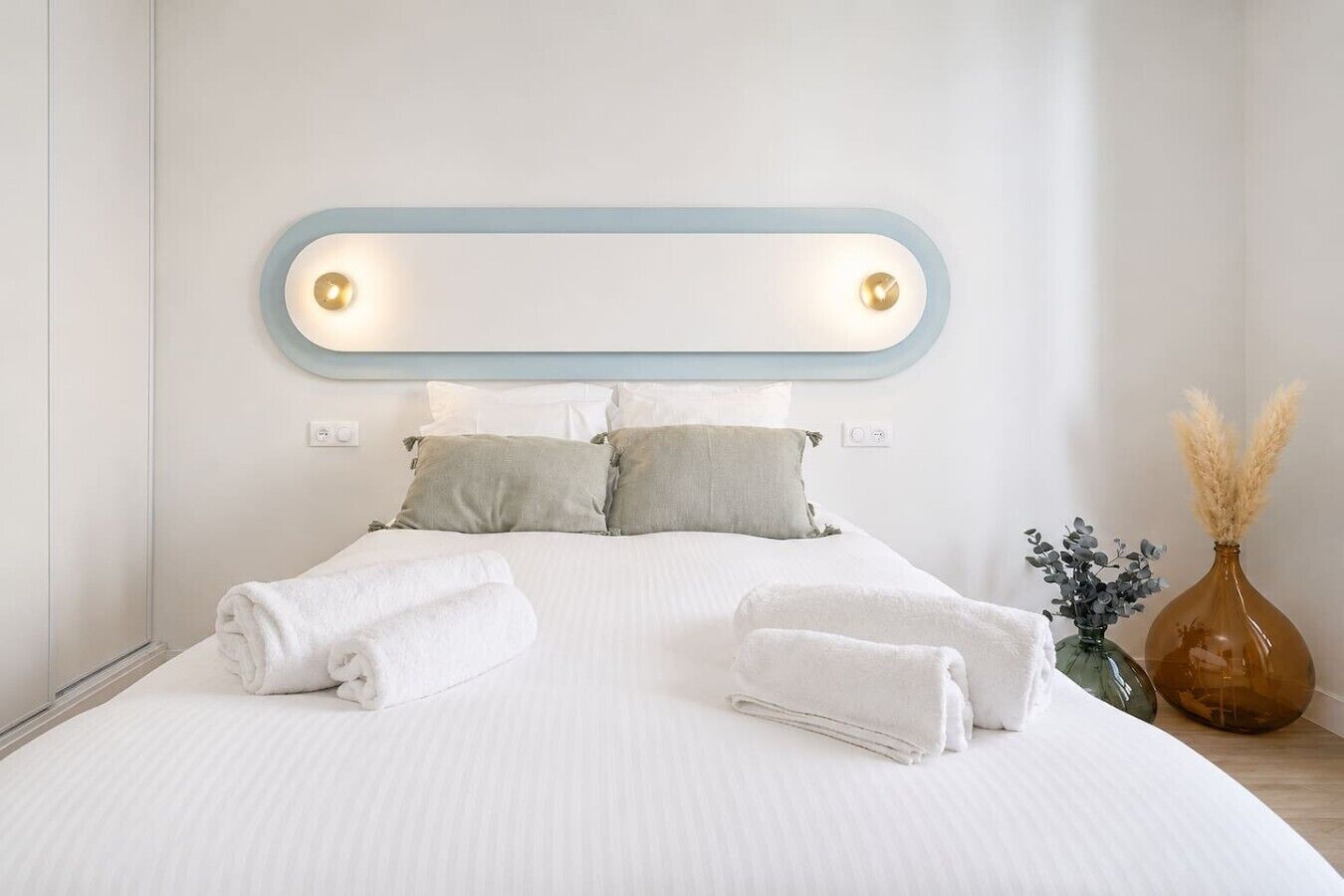 A neatly made bed with white linens and two pillows, a bedside table with a vase and a bottle, and a wall-mounted light fixture.