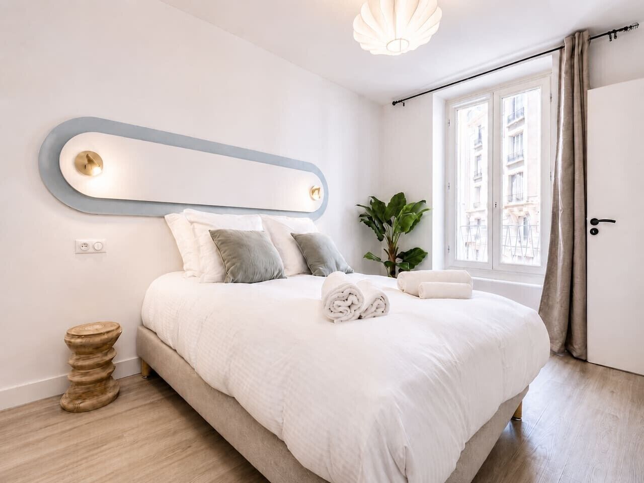 A modern bedroom with a large bed, a bedside table, a mirror, and a window with curtains.