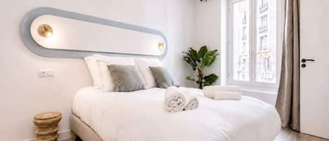 A modern bedroom with a large bed, a bedside table, a mirror, and a window with curtains.