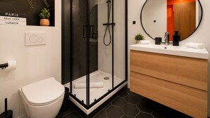 Et moderne badeværelse med en glasafskærmet brusekabine, et hvidt toilet, en trækommode og et rundt spejl.
