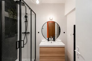 A modern bathroom with a glass shower enclosure, a round mirror with a wooden door, and a white sink with a wooden vanity. - Lavie Maison | Eiffel Nearby - Beaugrenelle (Paris)