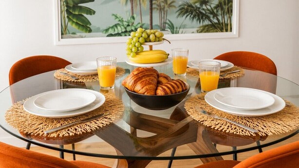 Et rundt glasbord med en hvid tallerken, en skål med croissanter, en druebundt, en banan og to glas appelsinjuice.