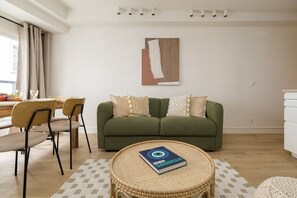 A living room with a green sofa, two patterned pillows, a round wicker table with a book, a dining area with chairs and a table, and a white wall with a painting. - Lavie Maison Cozy Flat in Parisian Passage (Paris)