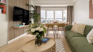 Una sala moderna con un sofá verde, un comedor con mesa para cuatro, un televisor de pantalla plana montado en un panel de madera, y un amplio ventanal con vistas a la ciudad.