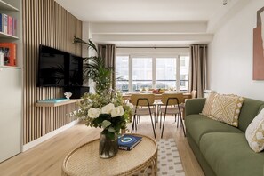 Ein modernes Wohnzimmer mit einem grünen Sofa, einem Essbereich mit Tisch für vier Personen, einem an einer Holzverkleidung angebrachten Flachbildfernseher und einem großen Fenster mit Blick auf die Stadt.