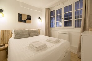 A neatly made bed with pillows and folded towels, a bedside table, a wall-mounted light, and a window with curtains. - Lavie Maison Cozy Flat in Parisian Passage (Paris)