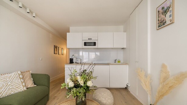 Un espacio habitable compacto con kitchenette, un sofá verde, un jarrón decorativo con flores y un cuadro colgado en la pared.