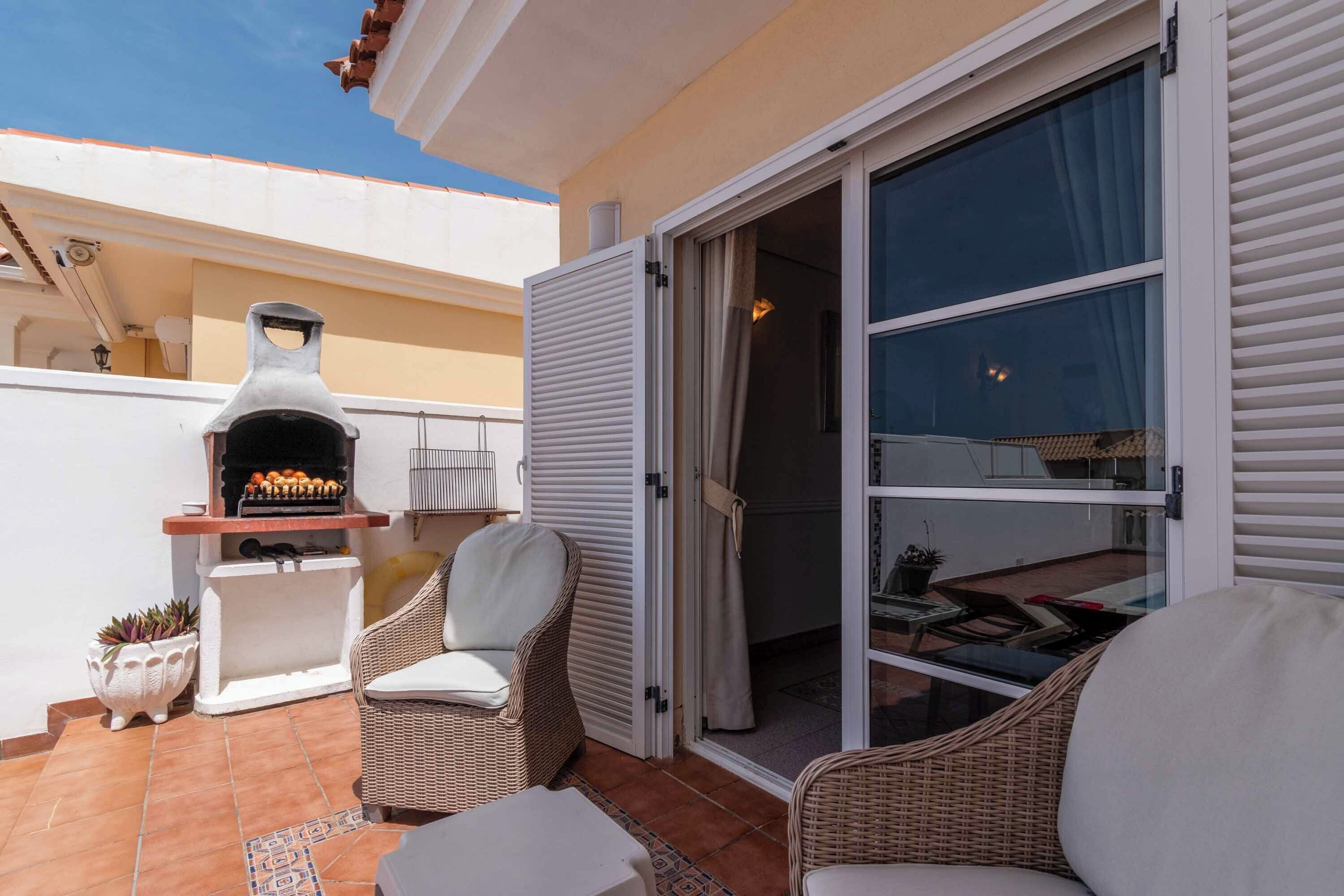 Un patio con una parrilla, mobiliario de mimbre y una puerta corrediza de vidrio que da a un espacio interior.