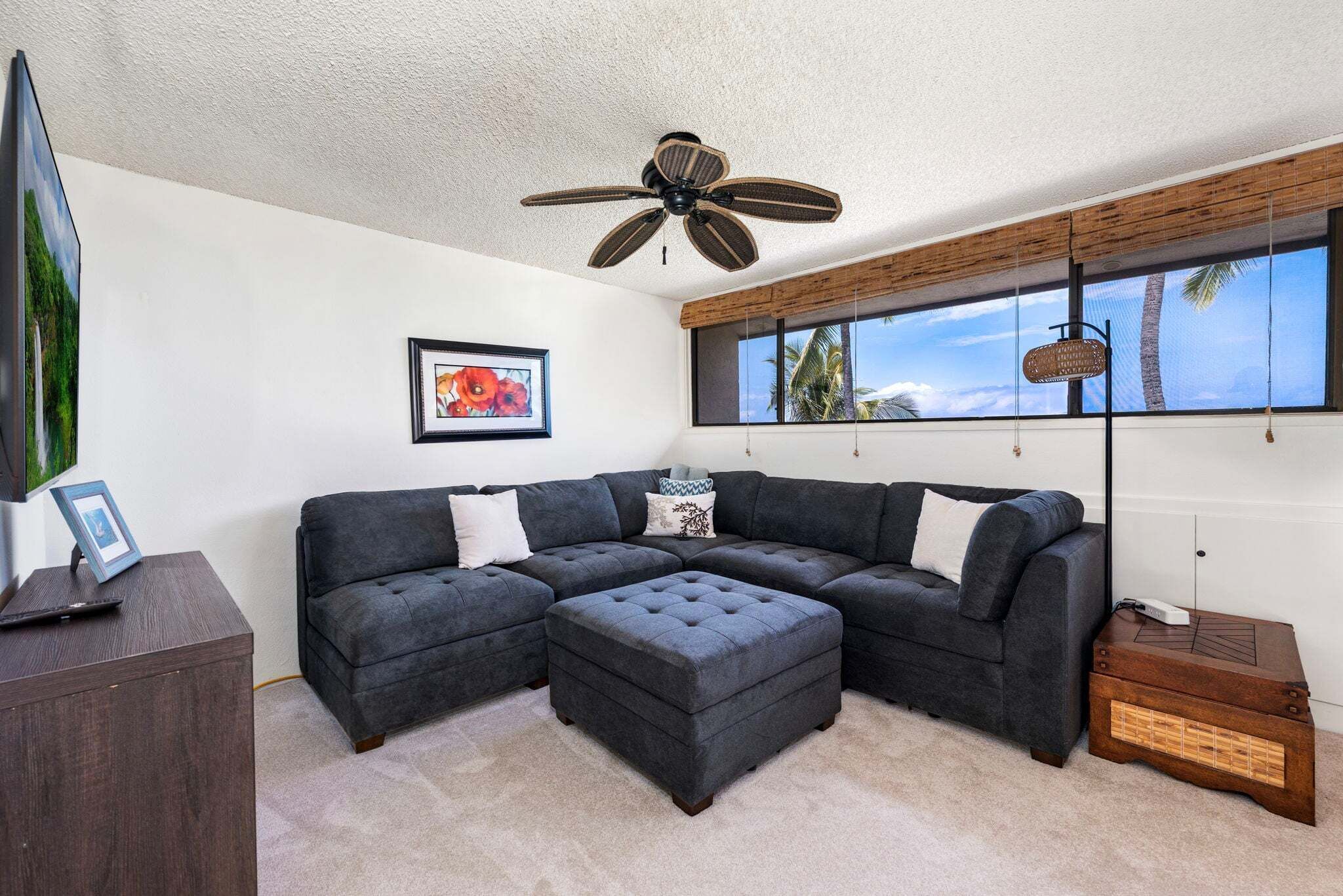 A living room with a sectional sofa, a tufted ottoman, a ceiling fan, a framed picture, and a window with a view of palm trees.
