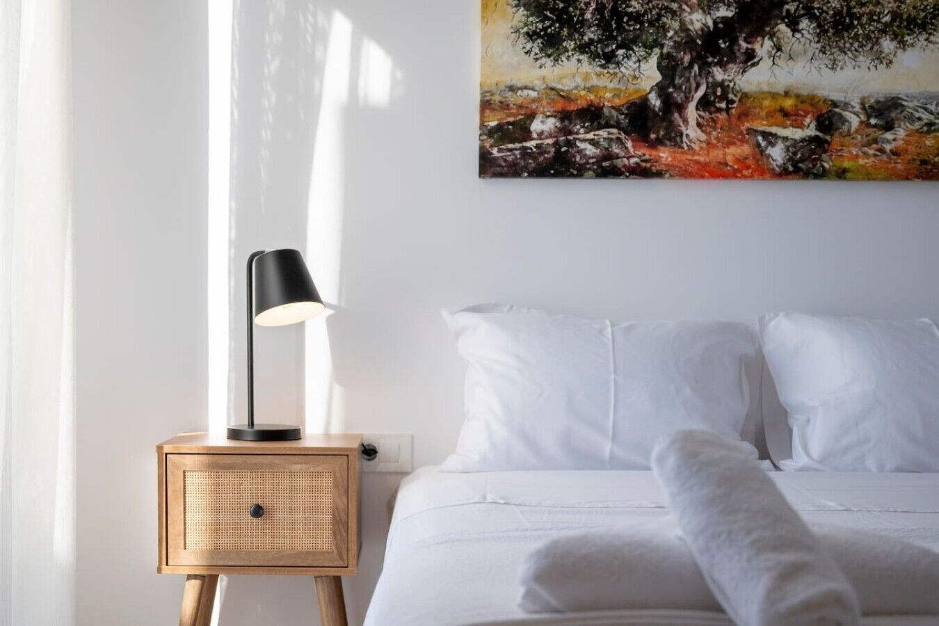 Ein Schlafzimmer mit einem Bett, weißen Kissen, einer schwarzen Lampe auf einem Nachttisch aus Holz und einem Baumgemälde an der Wand.