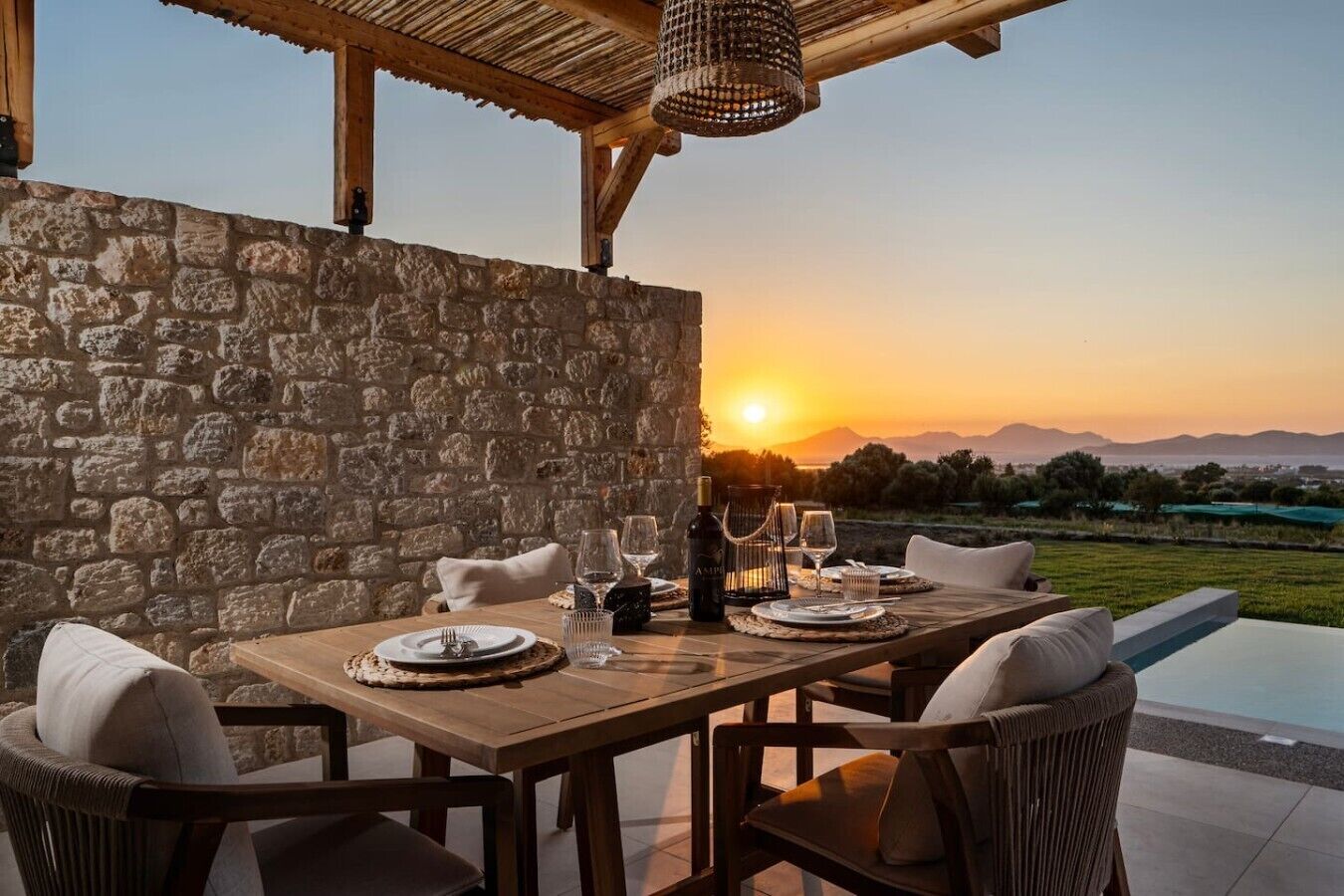 Un espace de restauration en plein air, agrémenté d’un mur en pierre, d’une table en bois et de chaises, offrant une vue sur une piscine et sur le coucher du soleil.