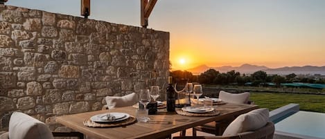An outdoor dining area with a stone wall, wooden table, and chairs, overlooking a pool and a sunset view.