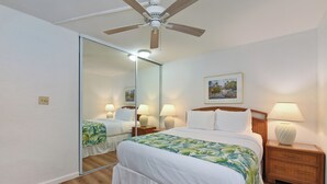 Ein Hotelzimmer mit einem Bett, einem Nachttisch, einer Lampe, einem Deckenventilator, einem Spiegel und einem gerahmten Bild an der Wand.