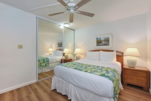 Ein Hotelzimmer mit einem Bett, einem Nachttisch, einer Lampe, einem Deckenventilator, einem Spiegel und einem gerahmten Bild an der Wand.