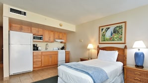 A hotel room with a bed, a nightstand, a lamp, a painting, a kitchen area with a refrigerator, microwave, and stove. - Sweet Studio with AC (Lahaina)