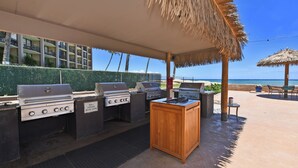 An outdoor barbecue area with multiple grills, a wooden table, and a view of the beach and hotel buildings. - Sweet Studio with AC (Lahaina)