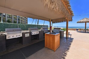 Ein Außenbereich mit Grillstationen, einem Holztisch und Blick auf Strand und Hotelgebäude.