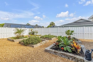 Ein Garten mit erhöhten Beeten, einem weißen Zaun und einer Solaranlage auf einem Dach.