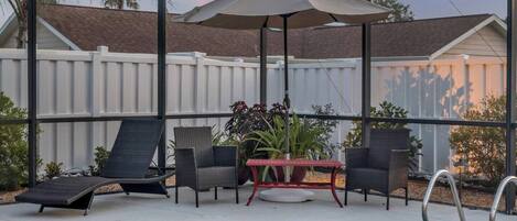 Un espace aménagé au bord de la piscine, doté d’une structure en verre, de deux chaises longues, d’une table ornée de plantes en pot et d’une piscine.