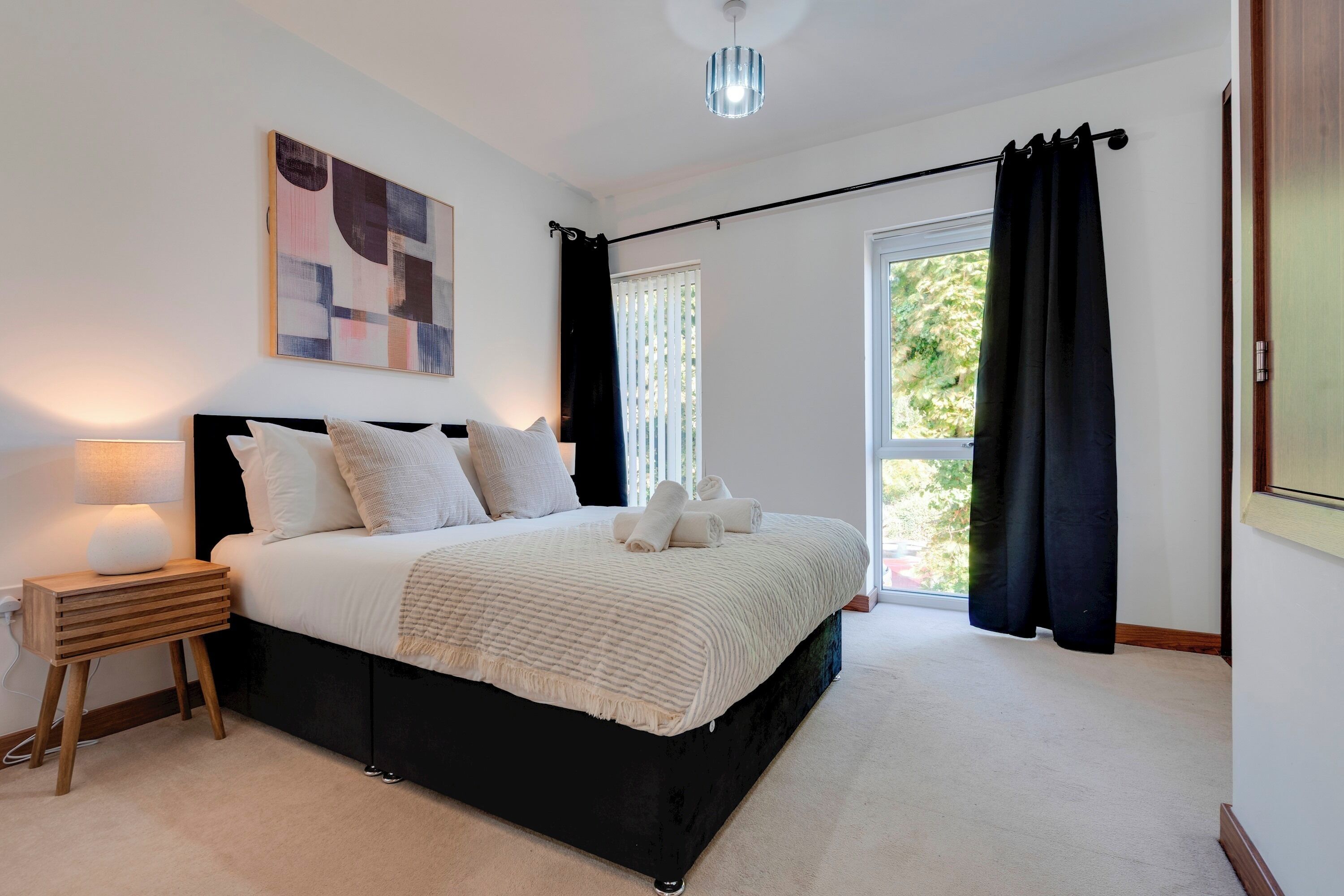 A bedroom with a bed, a nightstand, a lamp, a window with curtains, and a painting on the wall.