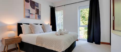 A bedroom with a bed, a nightstand, a lamp, a window with curtains, and a painting on the wall.