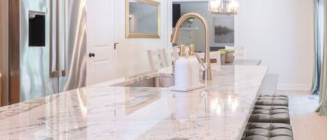 A modern kitchen with a marble countertop, black cushioned bar stools, and pendant lighting.