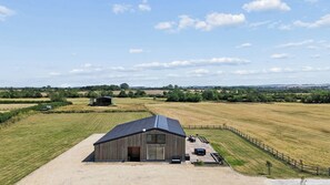 A large barn with a sloped roof, surrounded by open fields and a clear sky. - Secluded, contemporary barn in expansive gardens - Pass the Keys (Wantage)