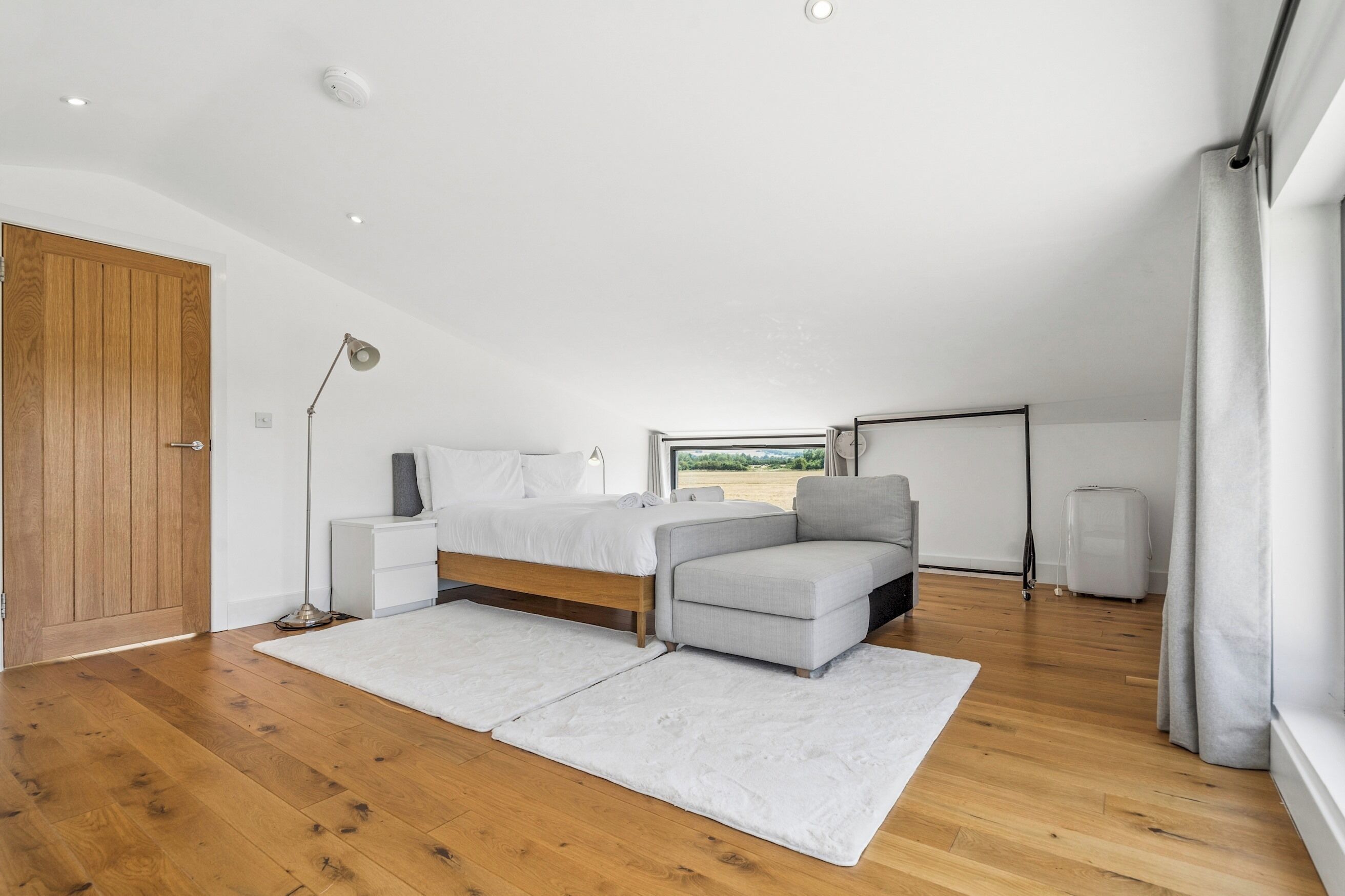 Un dormitorio con suelo de madera, una cama, un sillón gris, una mesita de noche, una lámpara y una ventana con vistas.