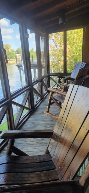 A wooden deck with a railing overlooking a body of water, featuring several wooden chairs and a bench.