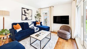 A modern living room with a blue sofa, a coffee table, a grey armchair, a TV, and a plant. - Large, Bright Westminster Apartment with Balcony (Greater London)