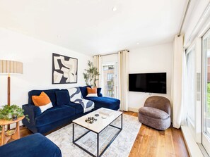 A modern living room with a blue sofa, a coffee table, a grey armchair, a TV, and a plant. - Large, Bright Westminster Apartment with Balcony (Greater London)
