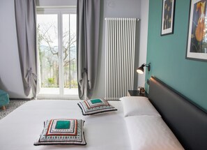 Habitació d'hotel amb un llit gran, dos coixins decoratius, una tauleta de nit amb una làmpada, una finestra amb cortines i un radiador.