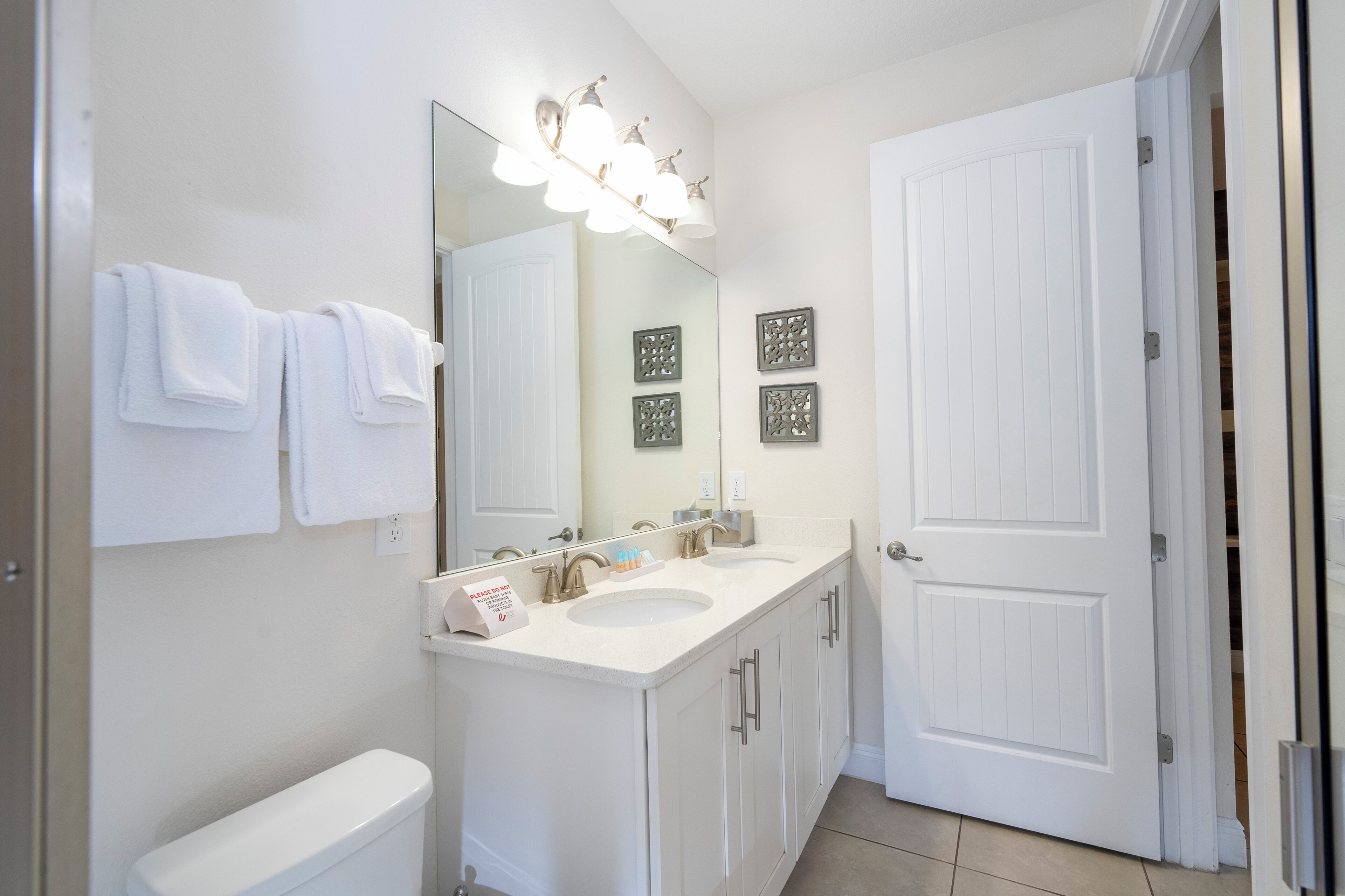 A bathroom with a white toilet, a sink, and a mirror with lights. There are towels hanging on the wall and a door with a handle.