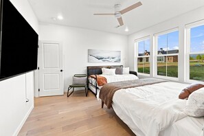 A bedroom with a bed, a chair, a television, a ceiling fan, and large windows with a view of houses. - 4 Peaks Post | 25min to Grand Targhee, Sleeps 18, Hot Tub, Views! (Tetonia)