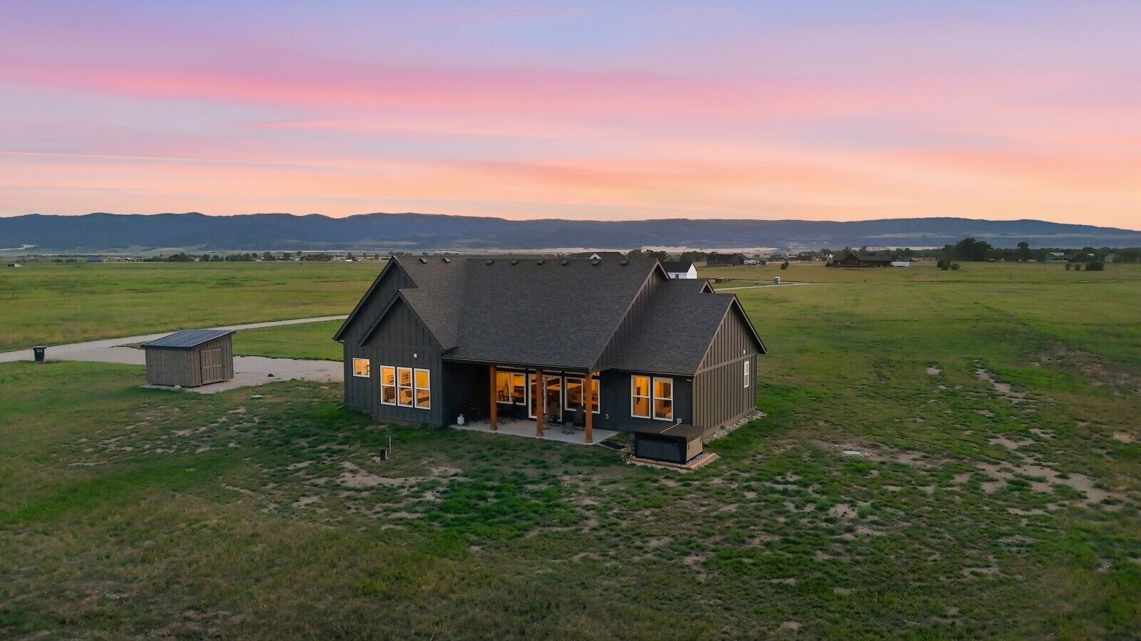 A single-story house with a dark roof and a front porch, surrounded by open fields and distant hills.