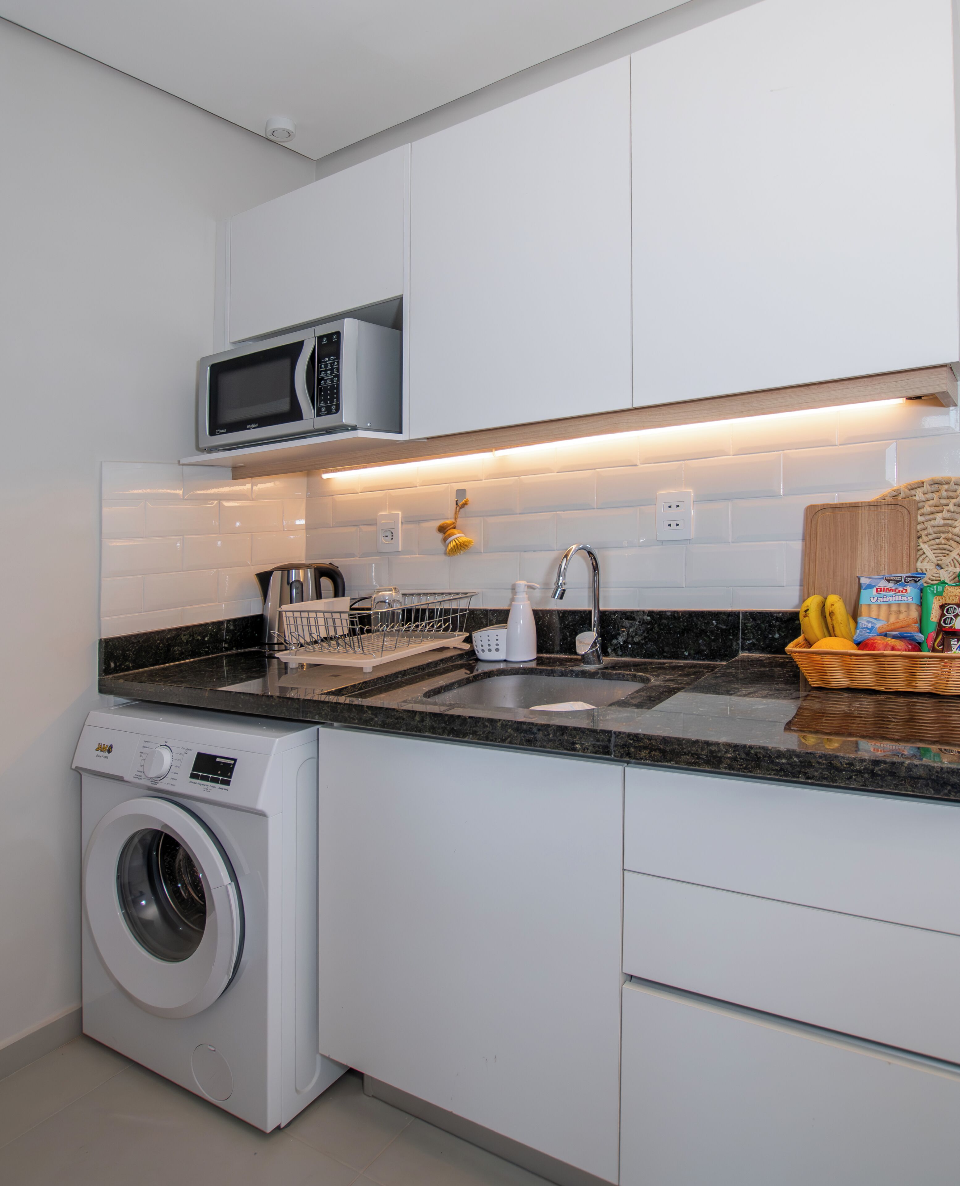 Una cocina moderna con lavadora integrada, microondas y una cesta de frutas sobre la encimera.