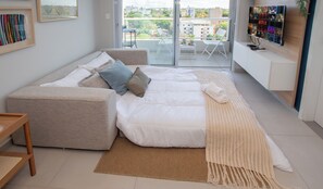 Ein modernes Wohnzimmer mit einer Couch, einem großen Bett, einem an der Wand befestigten Fernseher und Blick auf die Stadt durch das Fenster.