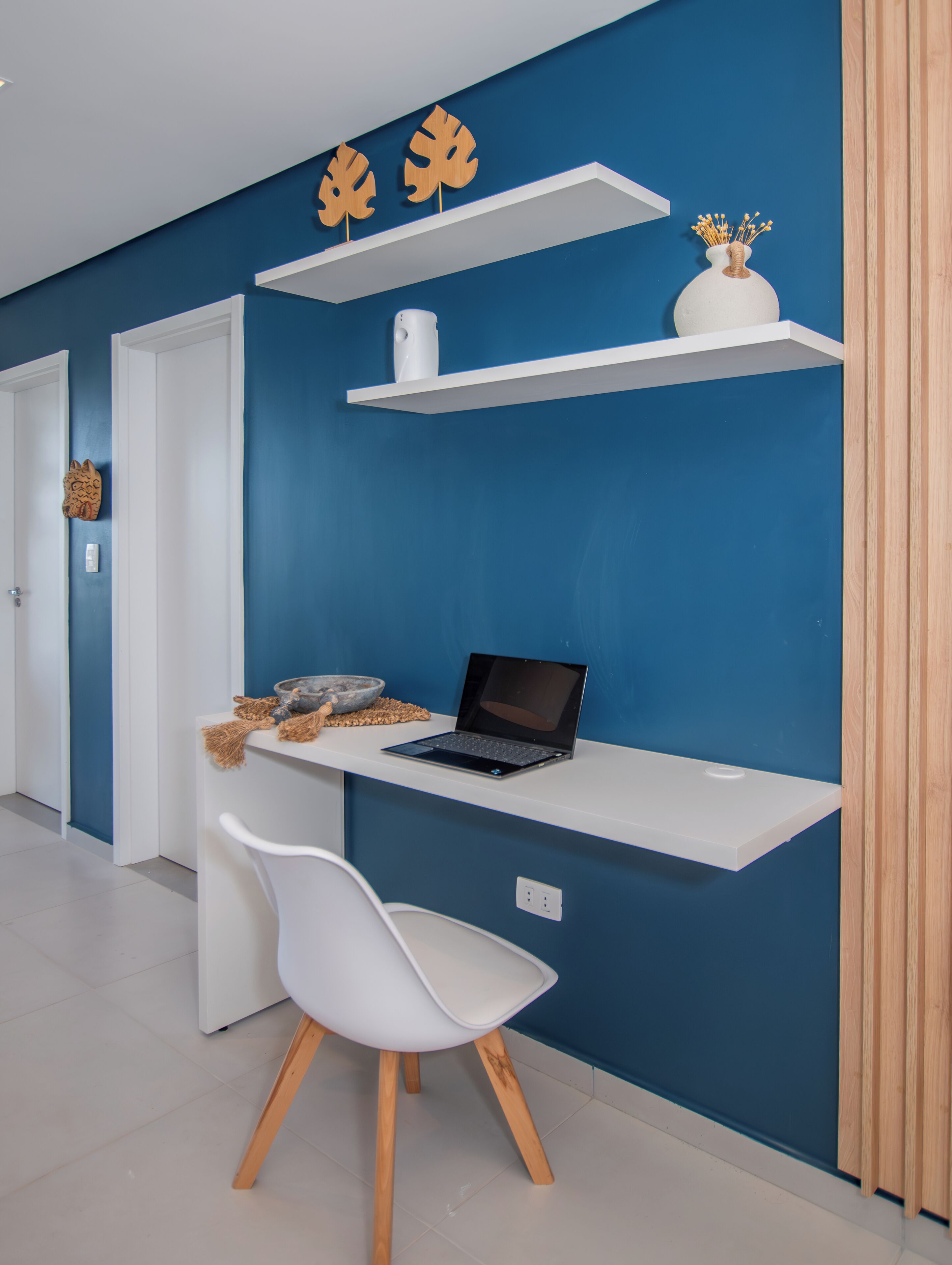 Une pièce moderne avec un mur bleu, des étagères blanches, un ordinateur portable, une chaise blanche et une porte en bois.