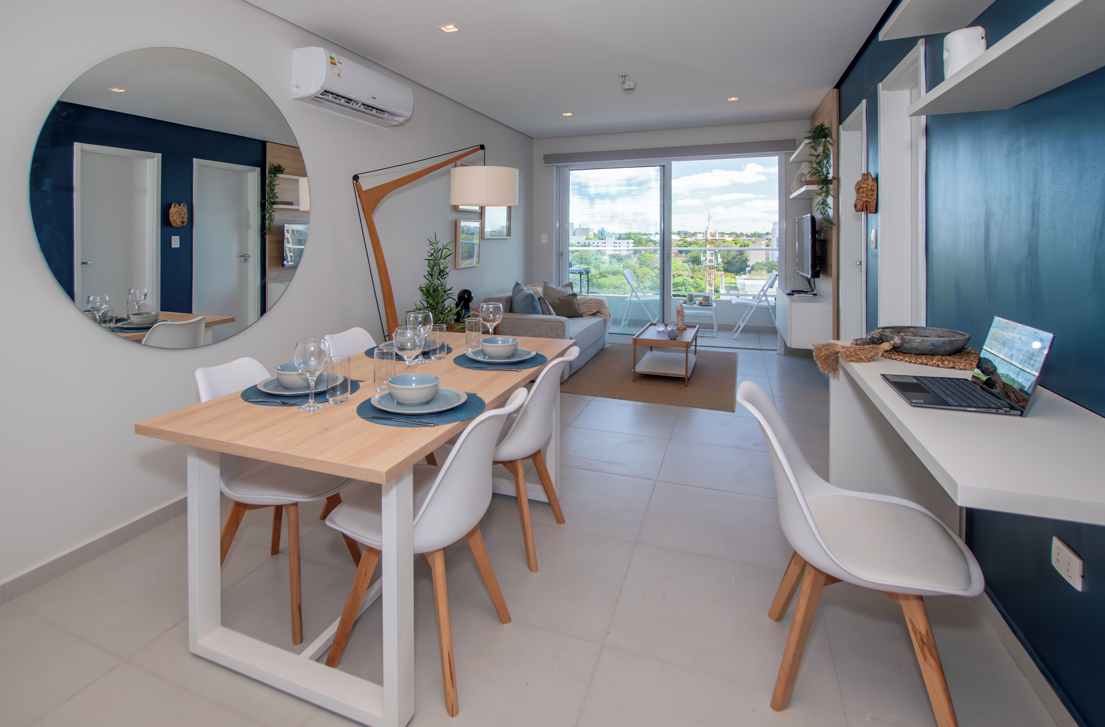 A modern dining area with a round mirror, a dining table set for six, and a sofa in the background.