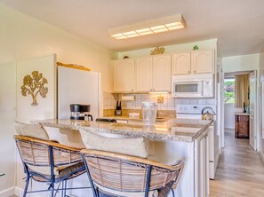 A kitchen with a central island, white cabinets, a microwave, and a refrigerator. There are two wicker chairs with cushions. - Mahina Surf Oceanfront Condo | Ground Floor | A/C (Lahaina)